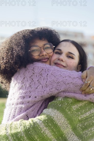 Two friends share a heartfelt hug, beaming with joy in a sunny outdoor setting. Both wear cozy knitted sweaters, radiating a sense of warmth and happiness
