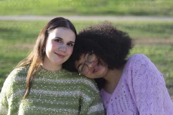 Two friends, one with long hair and the other with curly hair and glasses, wearing cozy sweaters, smile while enjoying a bright day outdoors in a green park setting