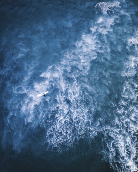 A dramatic aerial shot of a surfer on a white surfboard navigating through foamy blue waves in Portugal. Captures the energy and freedom of the ocean and the surfing lifestyle