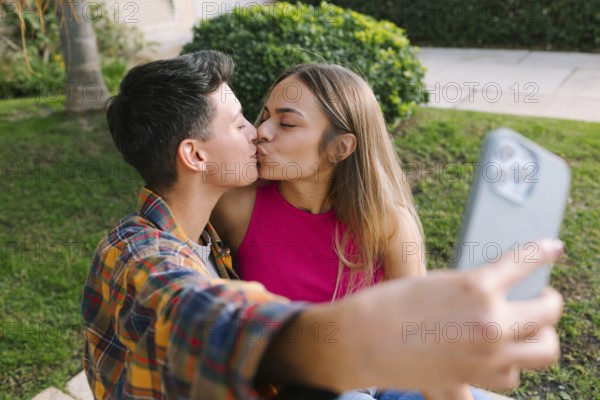 A loving lesbian couple shares a kiss while capturing the moment with a selfie. They sit in a green outdoor setting, expressing affection, happiness, and connection in a relaxed atmosphere