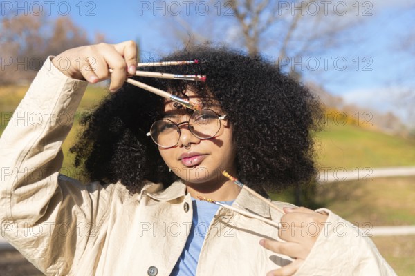 A creative young black artist holding several paintbrushes outdoors, surrounded by nature. The young woman expresses individuality and creativity, showcasing art in an inspiring environment