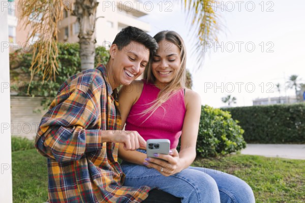 A happy lesbian couple smiles while looking at a phone screen in an outdoor setting. Their joyful expressions reflect love and connection, celebrating diversity