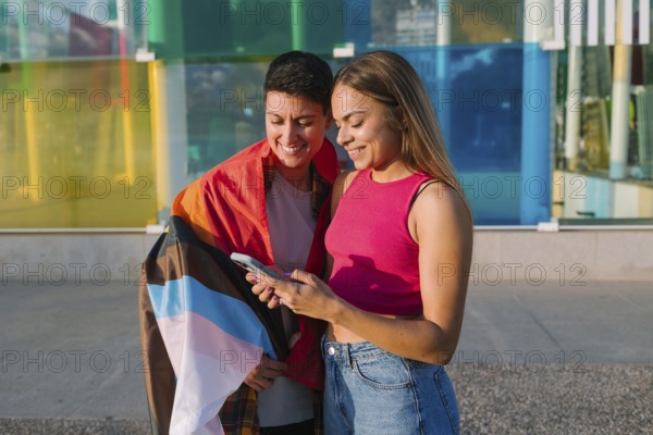 A happy lesbian couple looks at a smartphone together, smiling. One partner is wrapped in a Pride and Transgender flag, symbolizing LGBTQIA+ love, inclusivity, and self-expression in a vibrant setting