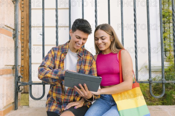 A happy lesbian couple sits together on a bench, smiling while looking at a tablet. One partner carries a Pride flag bag, symbolizing love, inclusivity, and LGBTQIA+ community pride