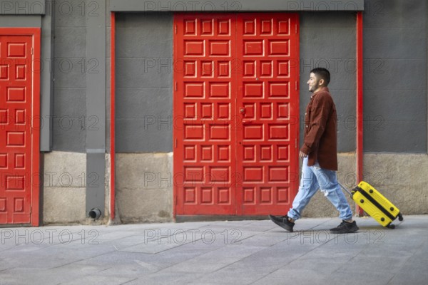 A latino transgender man walks confidently past vibrant red doors, pulling a bright yellow suitcase The urban scene reflects pride and journey