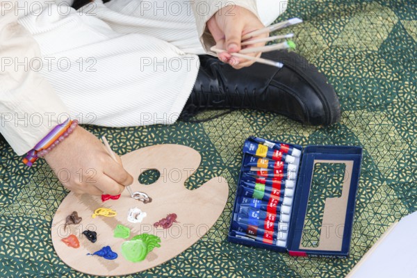 A close up of a young black woman preparing a wooden palette with colorful paint tubes and brushes on a patterned mat, showcasing creativity and artistic expression outdoors
