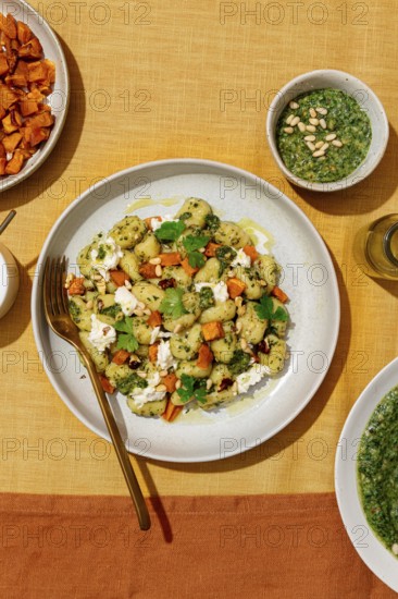 Top view of a colorful plate featuring potato gnocchi tossed with green pesto, garnished with parmesan, walnuts, and fresh herbs. Accompanied by roasted sweet potatoes and a bowl of pesto sauce on a vibrant yellow tablecloth