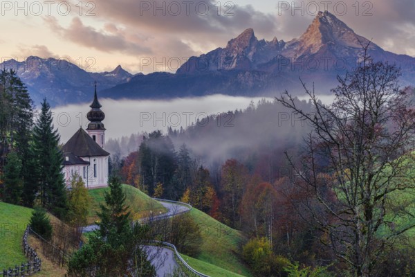 The picturesque Maria Gern Church is nestled among vibrant autumn foliage with the majestic Bavarian Alps towering in the background under a serene, misty sky