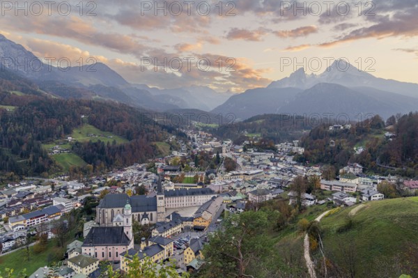 A stunning view of Berchtesgaden in the Bavarian Alps at sunset The charming alpine town, surrounded by lush hills and the iconic Watzmann mountain, glows under a vibrant evening sky