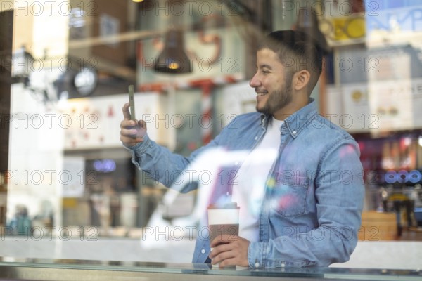 A Latino transgender man in a casual denim shirt smiles while holding a coffee and smartphone at a cafe Capturing diversity and authenticity in everyday moments