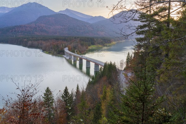 A scenic view of the Faller-Klamm Bridge spanning a tranquil lake in the Bavarian Alps Surrounded by autumn foliage and mountain peaks, this picturesque scene captures serene beauty