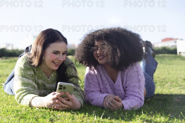 Two friends lie on grass, smiling and enjoying their time while looking at a smartphone in a sunny park, capturing a moment of technology and friendship