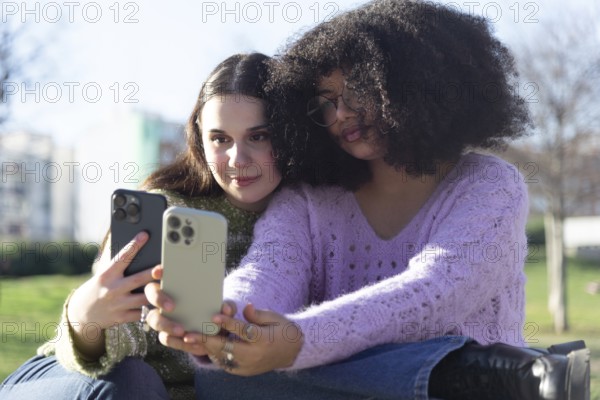 Two friends sitting on grass, taking selfies with their smartphones. They are enjoying a sunny day outdoors, wearing casual sweaters