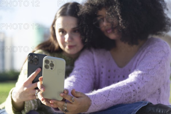 Two friends are sitting outdoors, capturing a selfie with smartphones. They are enjoying a sunny day in a park, sharing a moment of connection and joy
