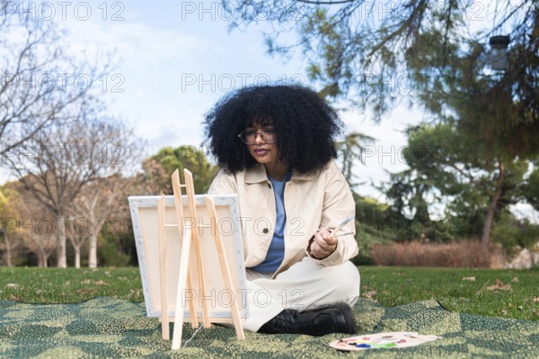Young black woman with curly hair immersed in painting outdoors, capturing a scenic day in nature. Surrounded by trees, she sits on a patterned blanket with a palette