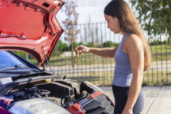 Young woman inspects a car engine on a sunny day The red car's hood is open, parked on a roadside She holds a dipstick, emphasizing post-accident vehicle maintenance