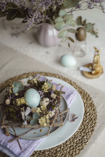 Top view of an Easter table setting featuring a plate with a rustic nest of twigs and naturally dyed eggs, surrounded by floral decorations and subtle tableware accents under soft, natural lighting