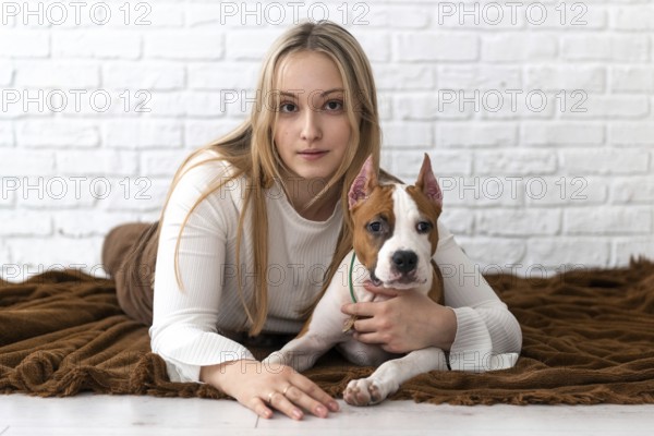 A young woman with blonde hair lounges on a brown blanket, affectionately cuddling her white and brown Staffordshire Terrier puppy dog in a warmly lit room with a white brick background