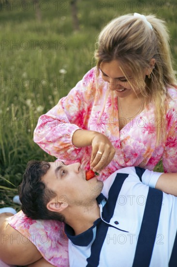 A loving couple shares a sweet moment outdoors as the woman playfully feeds her partner a strawberry. Their smiles and relaxed posture capture romance, joy, and togetherness in nature
