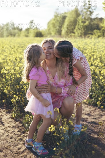 A joyful mother smiles as her daughters kiss her on both cheeks, sharing a tender moment in a beautiful flower field. The golden sunlight highlights their love and family bond