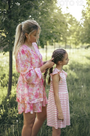 A loving mother gently braids her daughter's hair in a sunlit park. The tender moment captures care, connection, and the beauty of their mother-daughter bond in nature