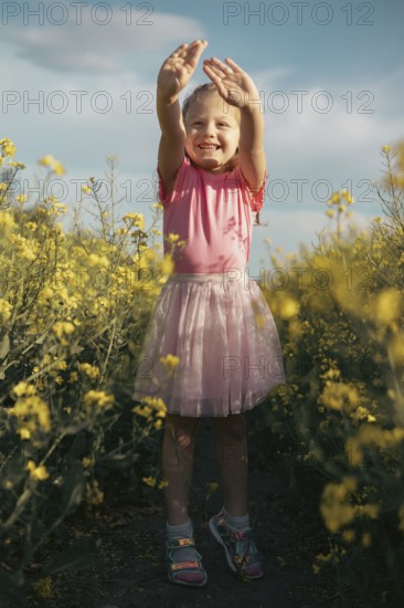 A joyful young girl in a pink outfit lifts her hands towards the sky while standing in a sunlit yellow flower field Her happiness and energy radiate a carefree childhood spirit