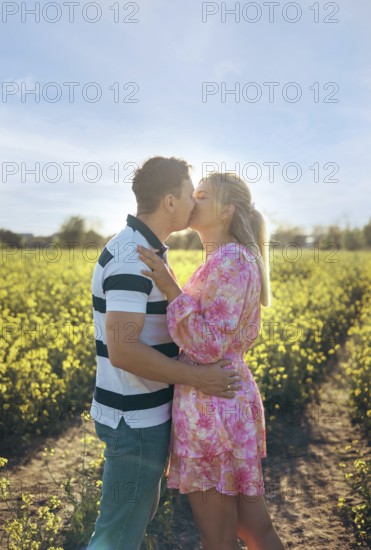 A loving couple embraces and shares a kiss in a golden flower field during sunset. The warm sunlight enhances the romance, creating a beautiful and intimate outdoor moment