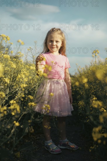 A young girl in a pink dress stands among vibrant yellow flowers, gently holding a bloom as she explores nature. Her wonder and curiosity capture the magic of childhood