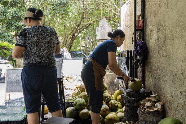 Two women prepare coconuts at a street stall, using a machete to open them. Fresh coconuts pile in the background, offering a glimpse into tropical street food culture