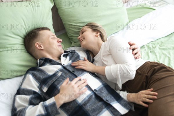 A young man and woman lie comfortably on a bed, sharing a close, intimate moment. They smile at each other, expressing affection and happiness in a cozy home setting