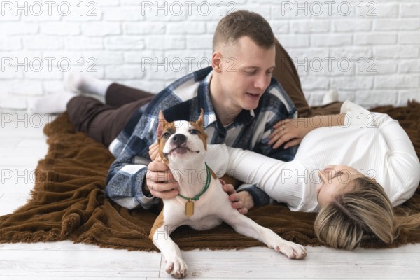 A young man and woman enjoy a cozy moment with their small white and tan dog on a brown blanket in a bright, white-walled room