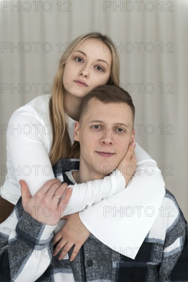 A young man sits as a woman embraces him from behind, both looking calm and affectionate at their cozy home setting