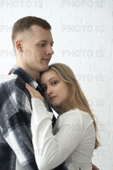 A young man and woman share a tender embrace in their home, expressing closeness and comfort. Both individuals are displaying a gentle affection in a bright, softly-lit room