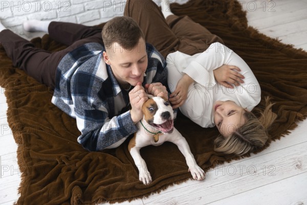 A man and a woman enjoy a playful moment with their dog, reclining on a plush brown blanket in a warmly lit room. Their expressions of joy and contentment mark a perfect family moment