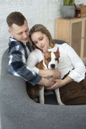 A young man and woman sit closely on a couch, affectionately holding a white and brown Staffordshire Terrier puppy dog, creating a warm, intimate scene of family and pet bonding in a cozy home environment