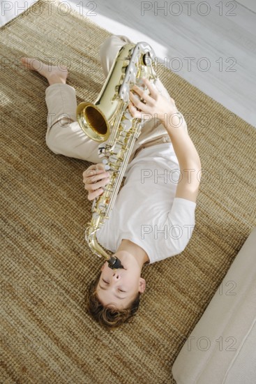 Young boy lying comfortably on a woven carpet and playing a saxophone. The cozy atmosphere emphasizes relaxation and the soothing pleasure of making music at home