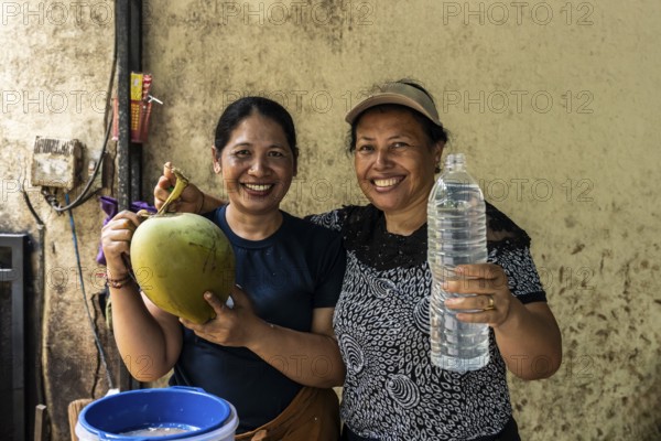Two smiling asian women showcase a freshly opened coconut and a bottle of coconut water, highlighting its refreshing taste. A perfect moment in a rustic setting