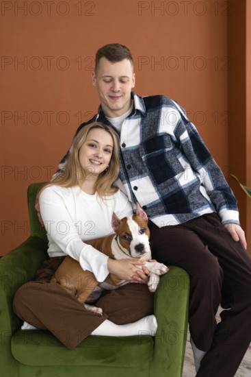 A young Caucasian man and woman smiling, seated comfortably in an armchair with their white and brown dog, in a well-lit room with orange walls