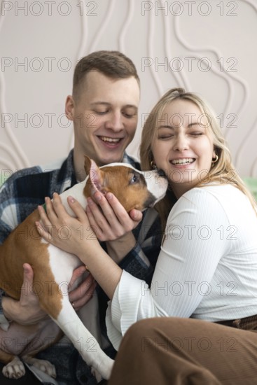 A cheerful young couple is seen enjoying a playful moment with their small brown and white dog in a cozy home setting