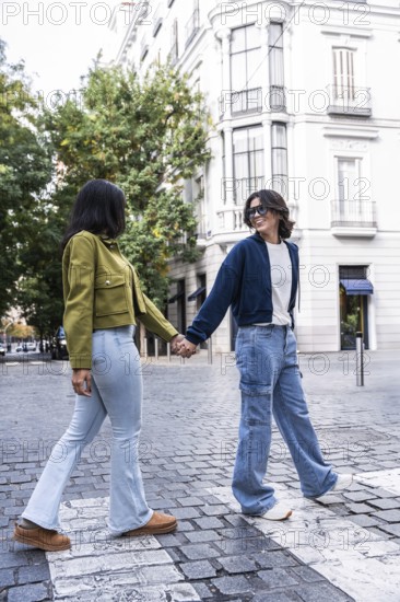 A lesbian couple holding hands while walking on a cobblestone street in the city, surrounded by architecture and greenery. They are smiling and enjoying the day