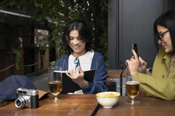 A lesbian couple shares a joyful moment at an outdoor cafe. One partner reads a book, and the other takes photos with a smartphone. Glasses of beer and a vintage camera sit on the table