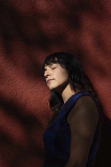 A Latin woman leans against a red wall with her eyes closed, basking in the sunlight. Shadows from nearby trees create an artistic and serene atmosphere, highlighting her peaceful expression