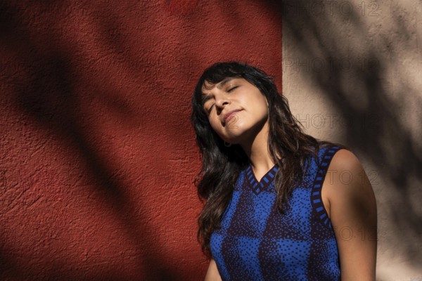 A Latin woman tilts her face toward the sunlight, eyes closed in a moment of relaxation. The warm tones of the red and beige wall, along with the shadows, create a serene and artistic atmosphere