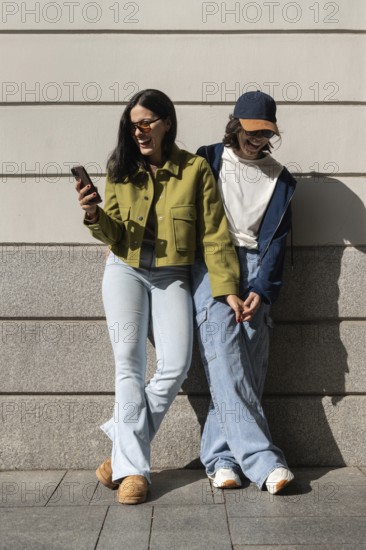 A joyful lesbian couple stands against a wall, holding hands and sharing laughter. The sunny day enhances their happiness as they engage in a moment of connection