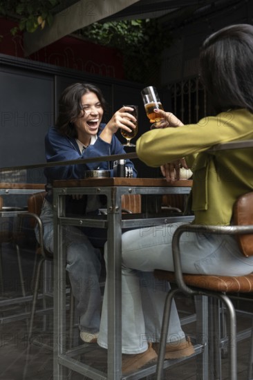 A lesbian couple joyfully toasts with glasses at a cozy cafe patio. They share a moment of laughter and connection, seated at a stylish table amidst urban greenery