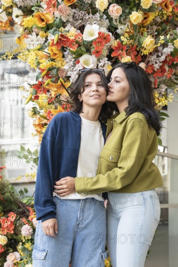 A lesbian couple shares a tender moment in front of a vibrant floral backdrop. Their closeness and happiness are highlighted by the bright, colorful flowers surrounding them