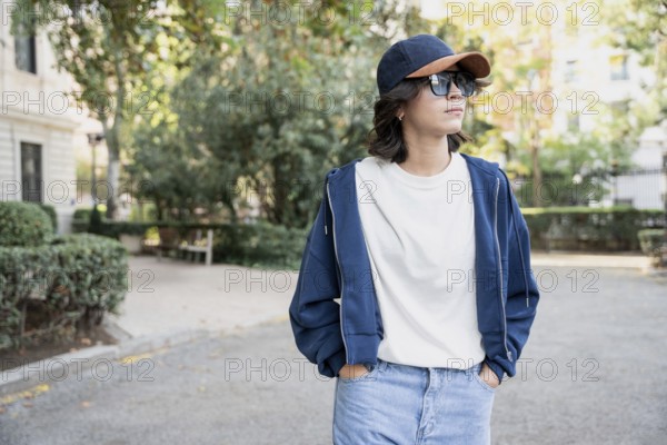 A latin woman wearing a navy cap, sunglasses, white shirt, blue hoodie, and jeans stands with hands in pockets, enjoying a sunny day in a park with trees and paths
