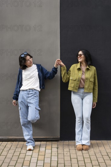 A joyful lesbian couple stands side by side, holding hands and smiling brightly while leaning against a stylish gray and black wall, showcasing love and happiness