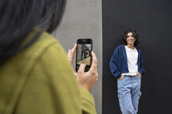 A lesbian couple enjoys a casual outdoor photoshoot. One woman wears denim and smiles against a dark wall, while the other captures the moment with her phone, wearing an olive jacket