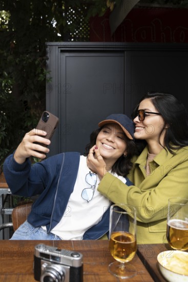 A joyful lesbian couple takes a selfie at a cozy outdoor cafe. They are smiling, surrounded by drinks and a camera, enjoying quality time on a sunny day